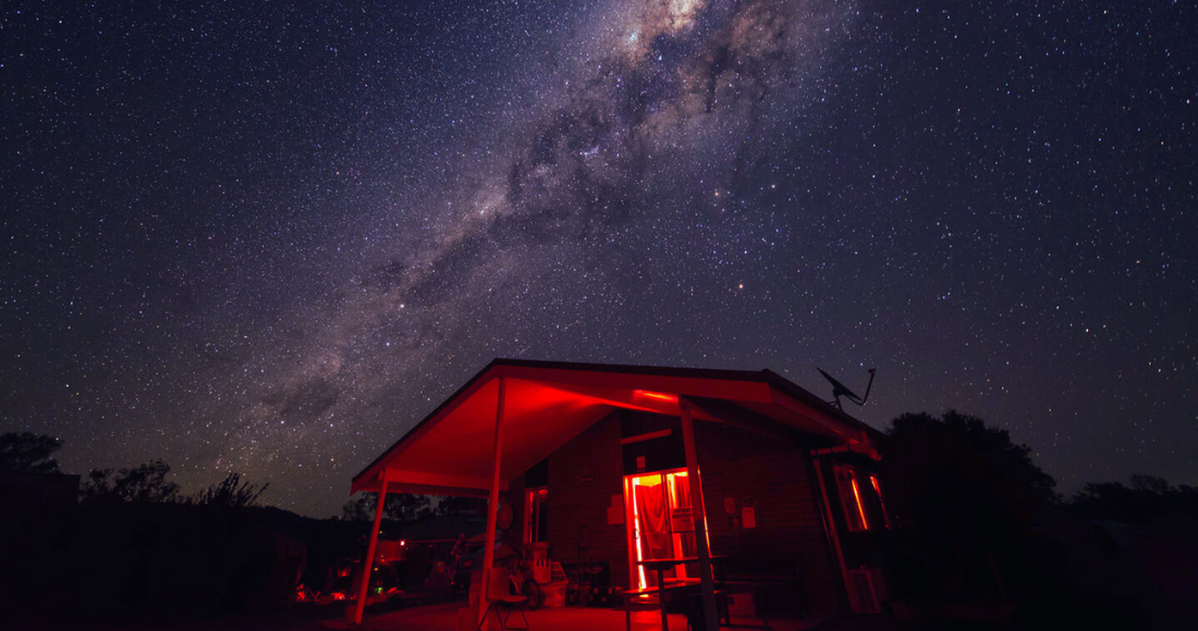 The 6 types of people you meet at star parties feature image, showing a small cabin or house bathed in red light and with a magnificent galaxy and sky above it