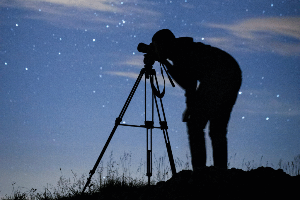 A silhouette of a man bending over a set of binoculars on a tripod at night, with a starry sky in the background.