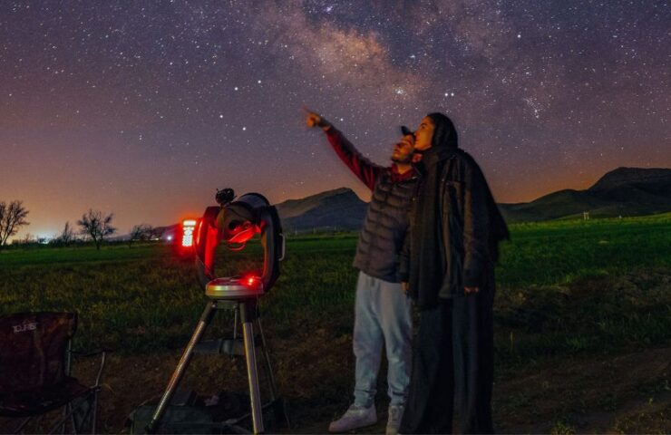 Two people stand in a field, oneof whom is pointing up at the sky. They're next to a telescope illuminated by a red light.
