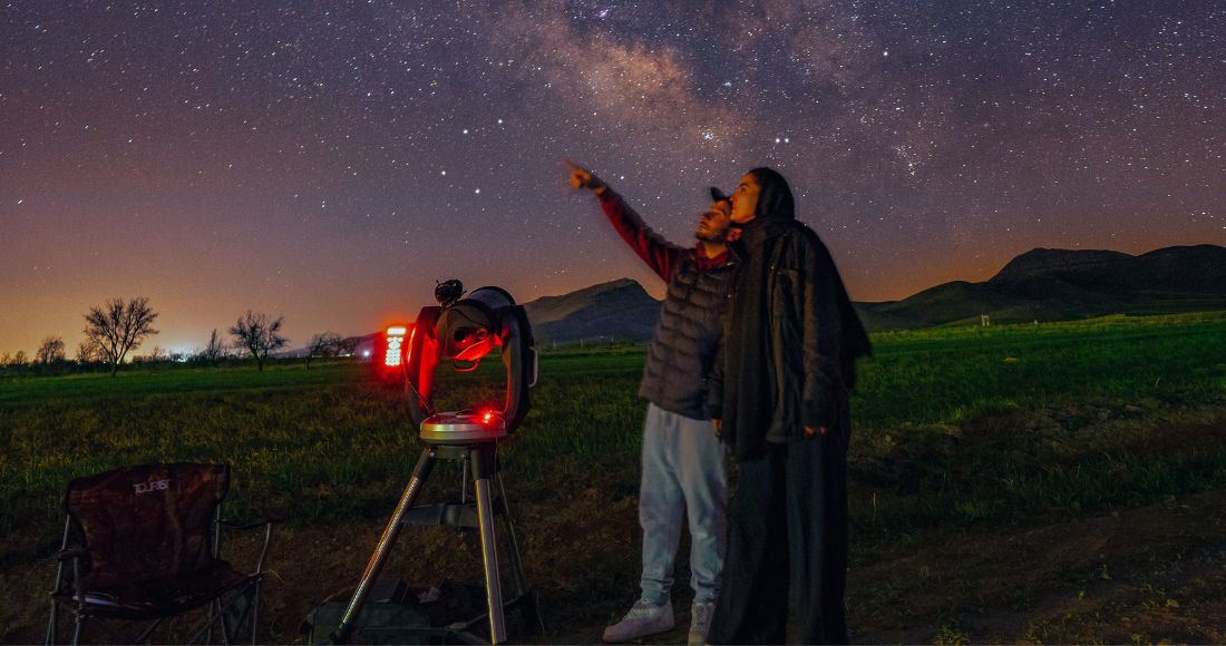 Two people stand in a field, oneof whom is pointing up at the sky. They're next to a telescope illuminated by a red light.