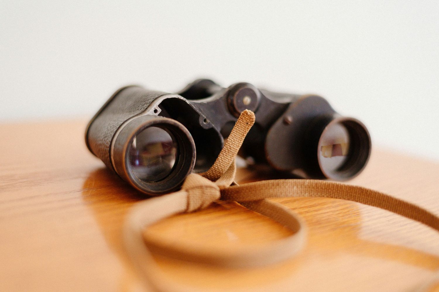 A close-up photo of a pair of older and slightly worn binoculars with a brown canvas strap lie on a light brown table