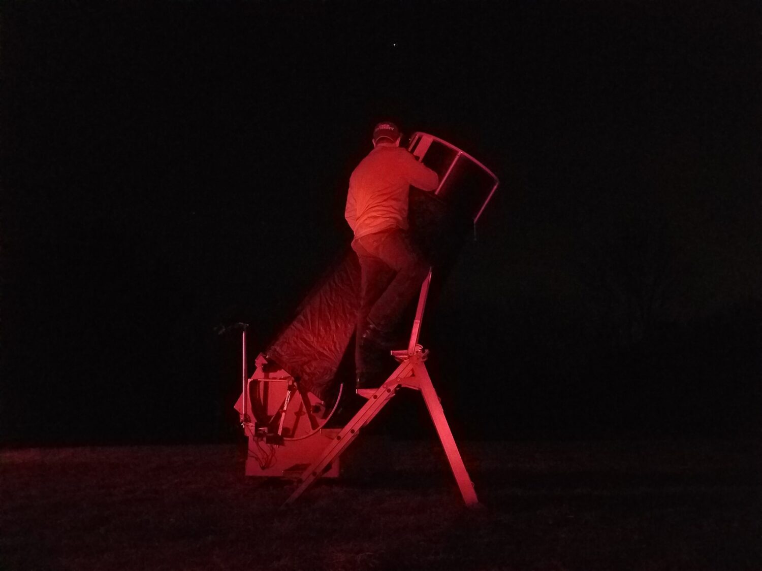 Josh standing on a ste-ladder to use his 25" Dobsonian at night, bathed in red light