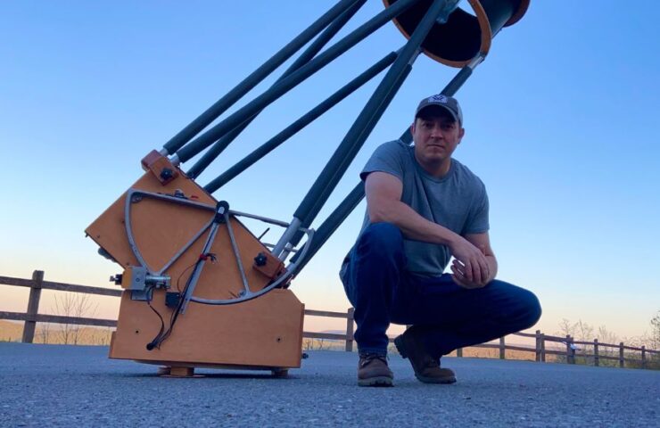 A man in jeans and a t-shirt kneels in front of a large dobsonian telescope