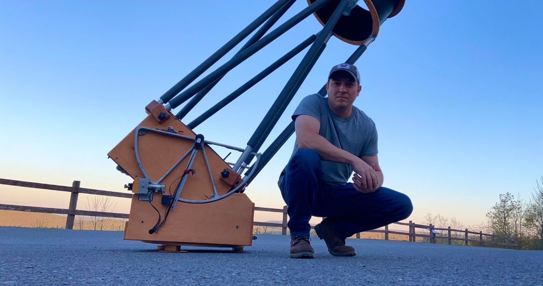 A man in jeans and a t-shirt kneels in front of a large dobsonian telescope