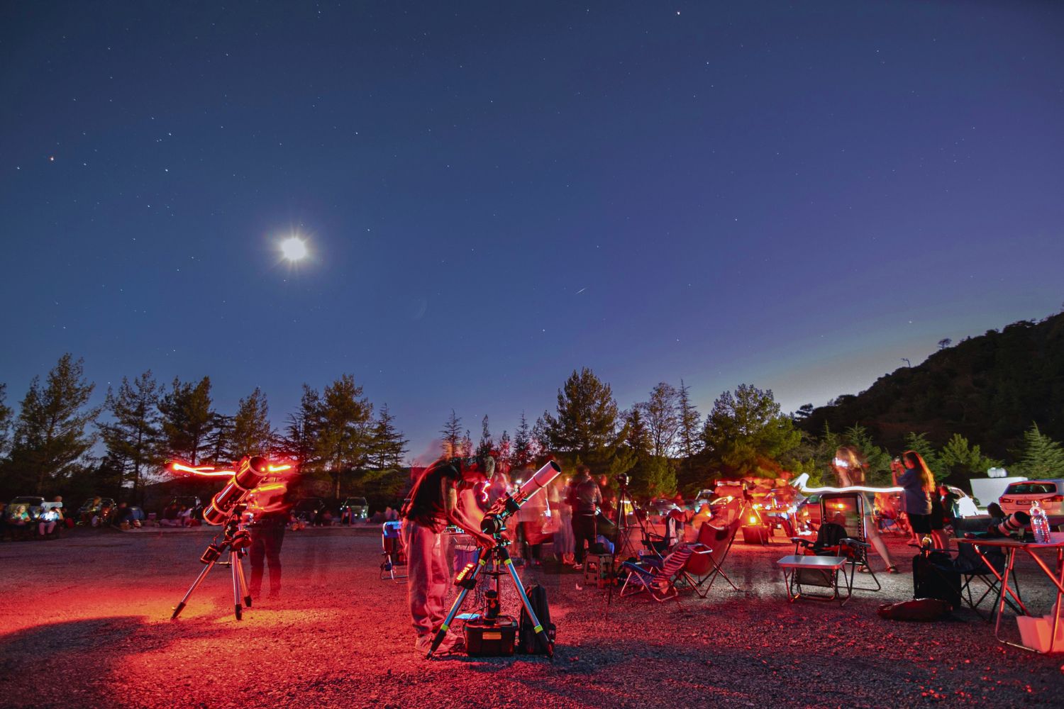 A stargazing night in Cyprus where dozens of people have set up telescopes, camp chairs and tables, and are lit up with red light as they use their telescopes.