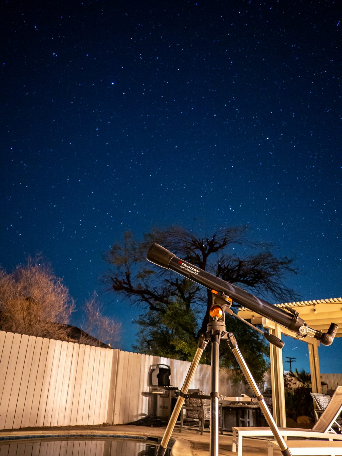 A small Celestron telescope stands next to a pool in a backyard, pointed at a night sky filled with stars